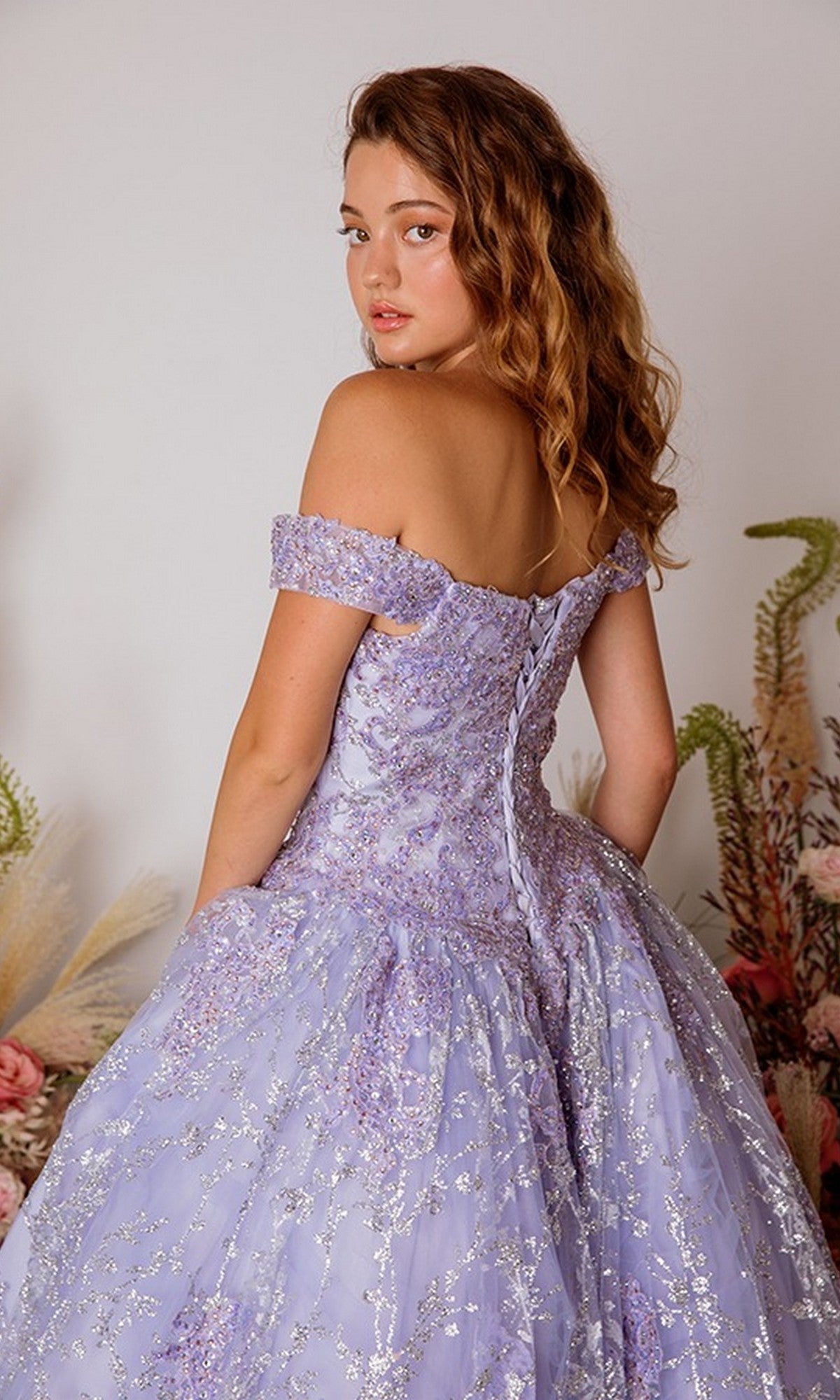 A young woman with wavy hair wears the Eureka 9909 Long Prom Dress, a lace off-the-shoulder lavender ballgown with silver embellishments, posing over her shoulder against a neutral backdrop with floral decor.